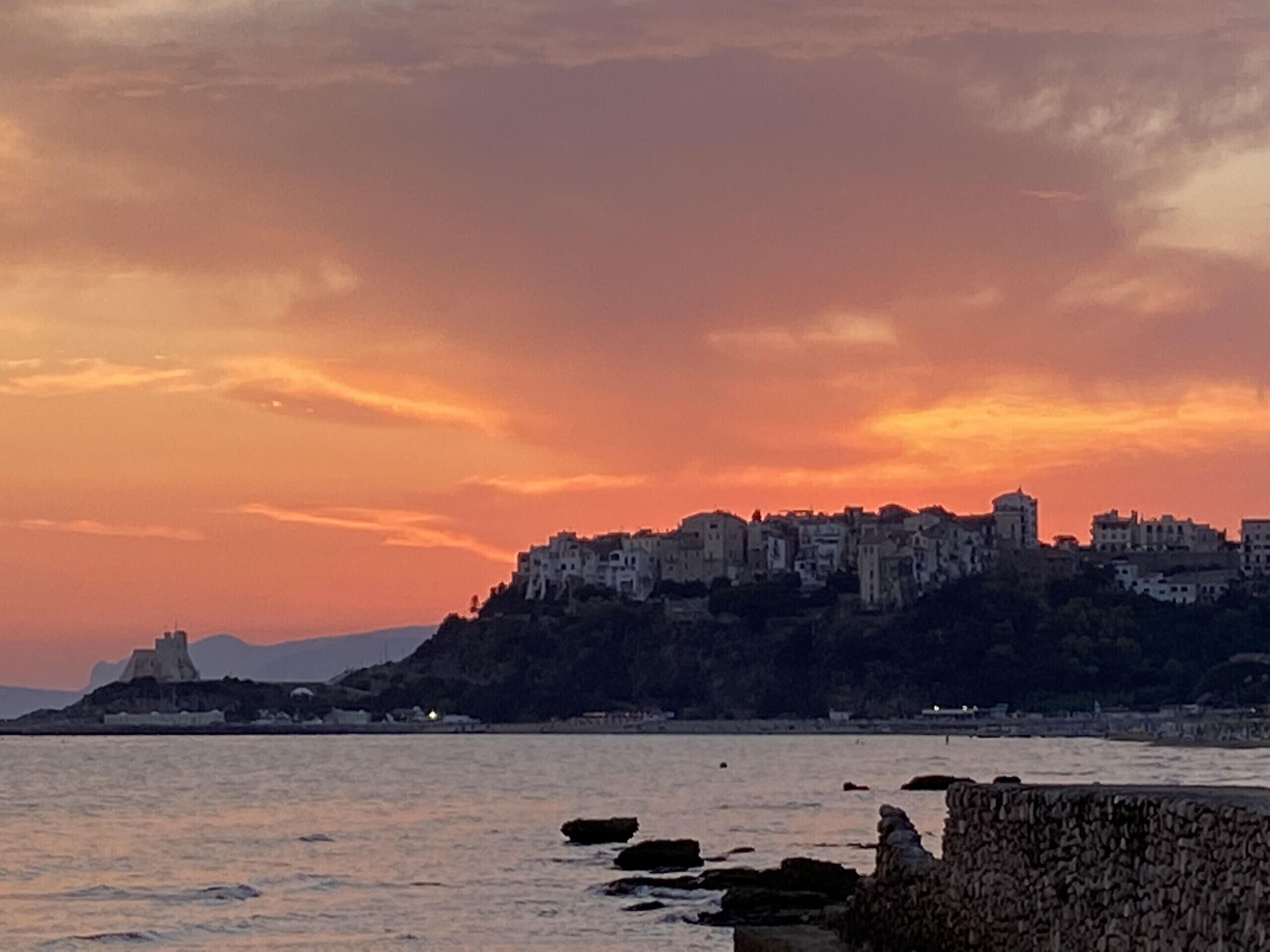 A coastal cityscape with a prominent hilltop building complex during sunset.