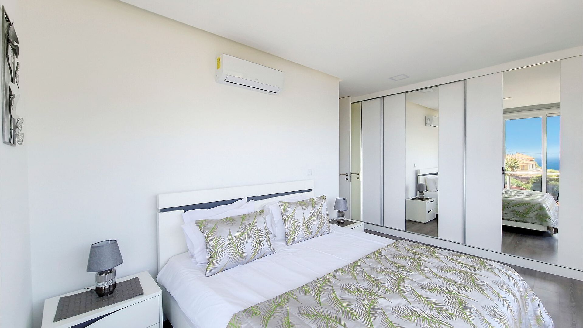 Un dormitorio moderno con una cama grande, una mesita de noche, una lámpara y vistas a un balcón con el paisaje urbano.