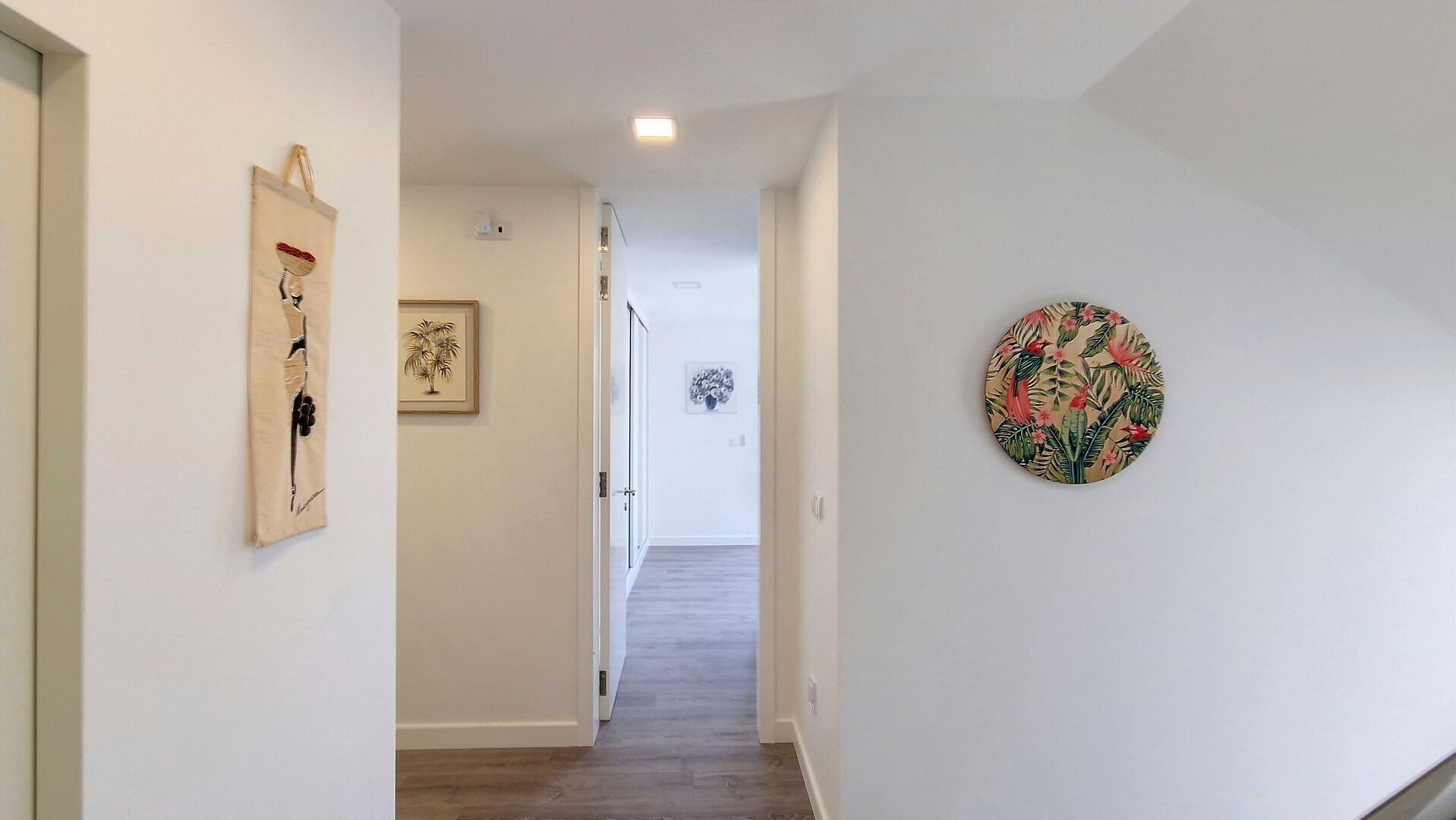 A hallway with a wooden floor, white walls, and artwork including a framed picture and a circular wall hanging.