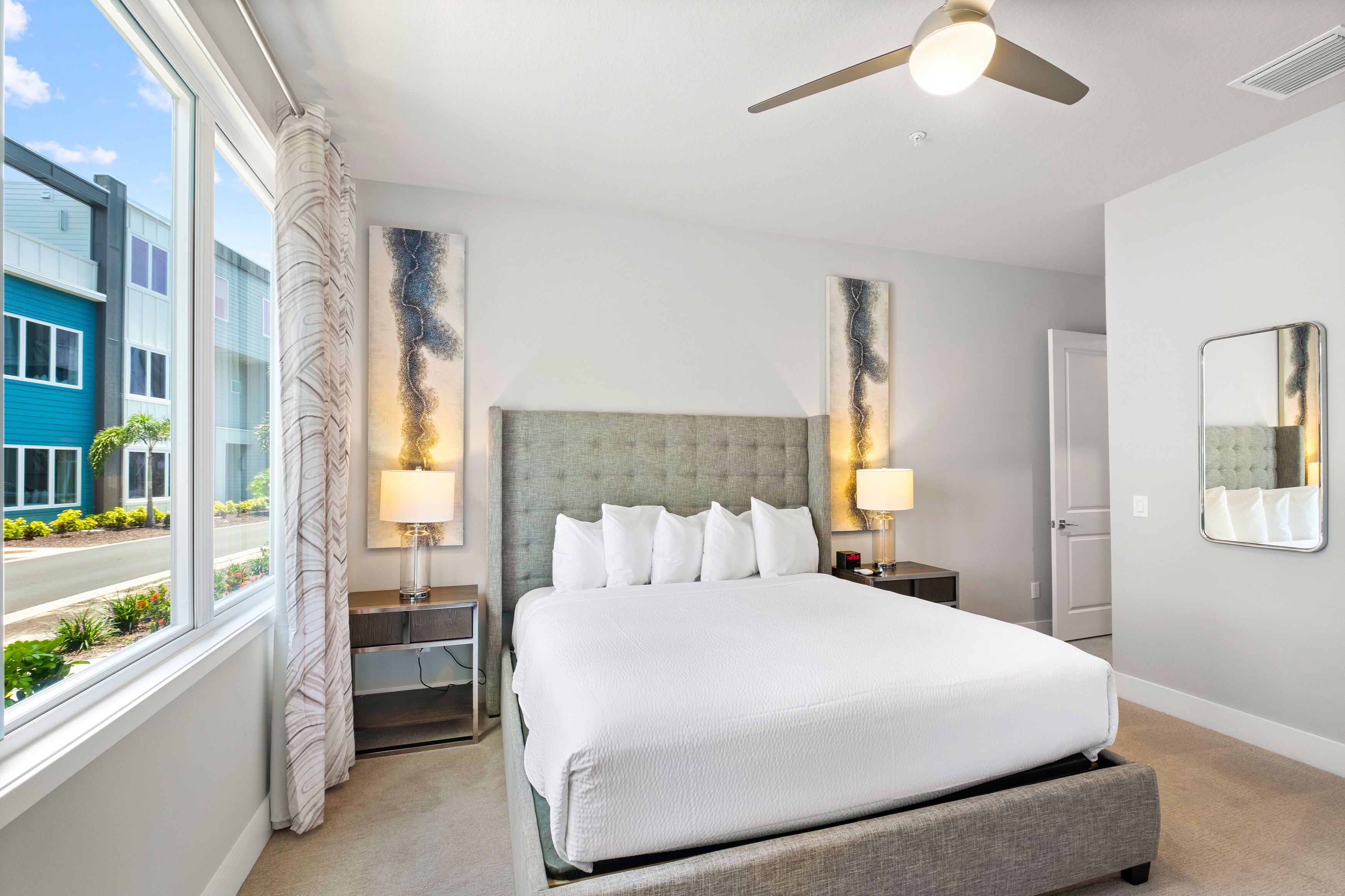 A hotel room with a large bed, two bedside tables, a ceiling fan, and a view of a building outside.