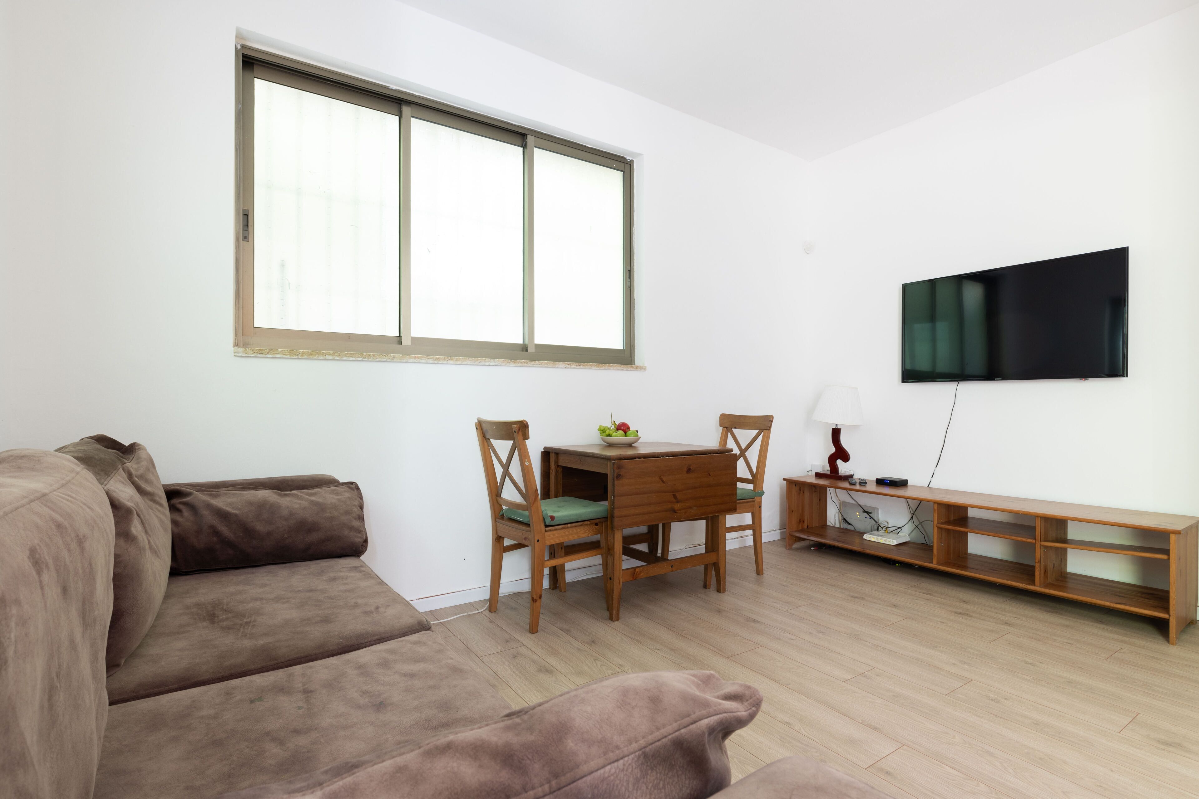 A living room with a brown sofa, a wooden dining table with two chairs, a TV mounted on the wall, and a wooden bench.