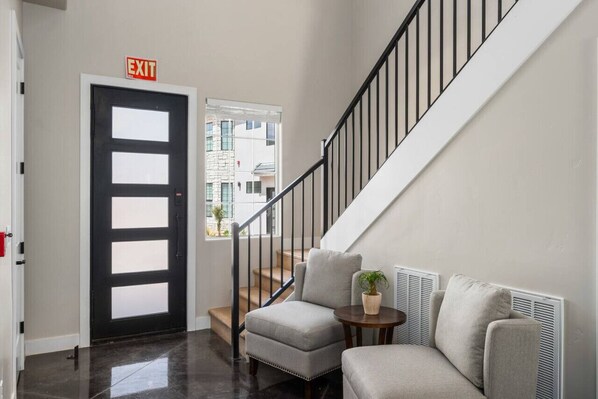 A modern interior with a staircase, a black door with an exit sign, a grey armchair, and a small table with a plant.