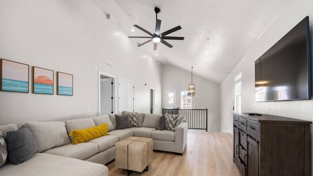 A spacious living area with a large L-shaped sofa, a wooden coffee table, and a dark wooden cabinet. A ceiling fan and a flat-screen TV are mounted on the wall.