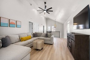 Un vaste espace de vie comprenant un grand canapé en forme de L, une table basse en bois et un meuble en bois foncé. Un ventilateur de plafond et un téléviseur à écran plat sont fixés au mur.