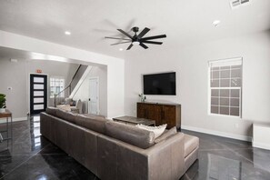 A modern living room with a large sectional sofa, a flat-screen TV mounted on the wall, and a ceiling fan.