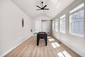 Um quarto amplo com mesa de futebol, ventilador de teto e grandes janelas que proporcionam luz natural.