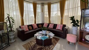 A living room with a dark brown sectional sofa, a coffee table, and a pink ottoman. There are two potted plants, one on the left and one on the right. The room has large windows with yellow curtains. - Pilot Hotel (Alaminos)