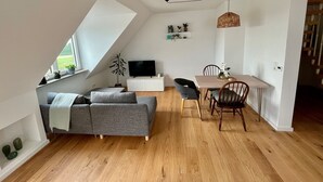 A modern loft apartment with a sloped ceiling, a dining area with a wooden table and chairs, a gray sofa, and a TV on a white cabinet.