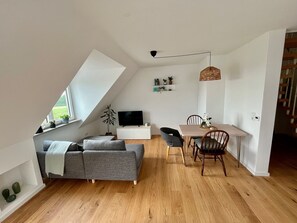 Un loft modern amb sostre inclinat, zona de menjador amb taula i cadires de fusta, sofà gris i televisió sobre un moble blanc.