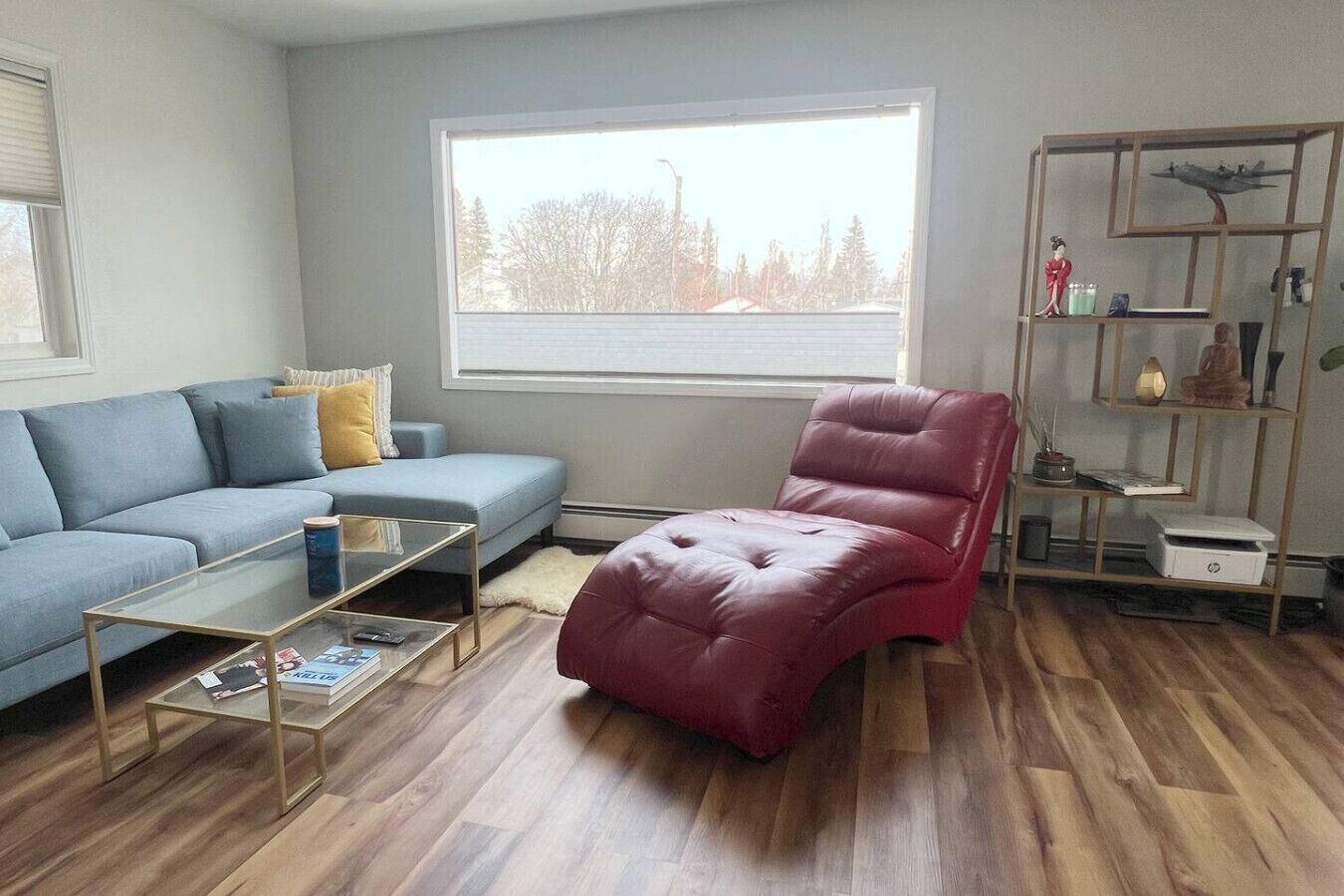 Una sala de estar con un sofá azul, un sillón rojo, una mesa de centro de vidrio y piso de madera.
