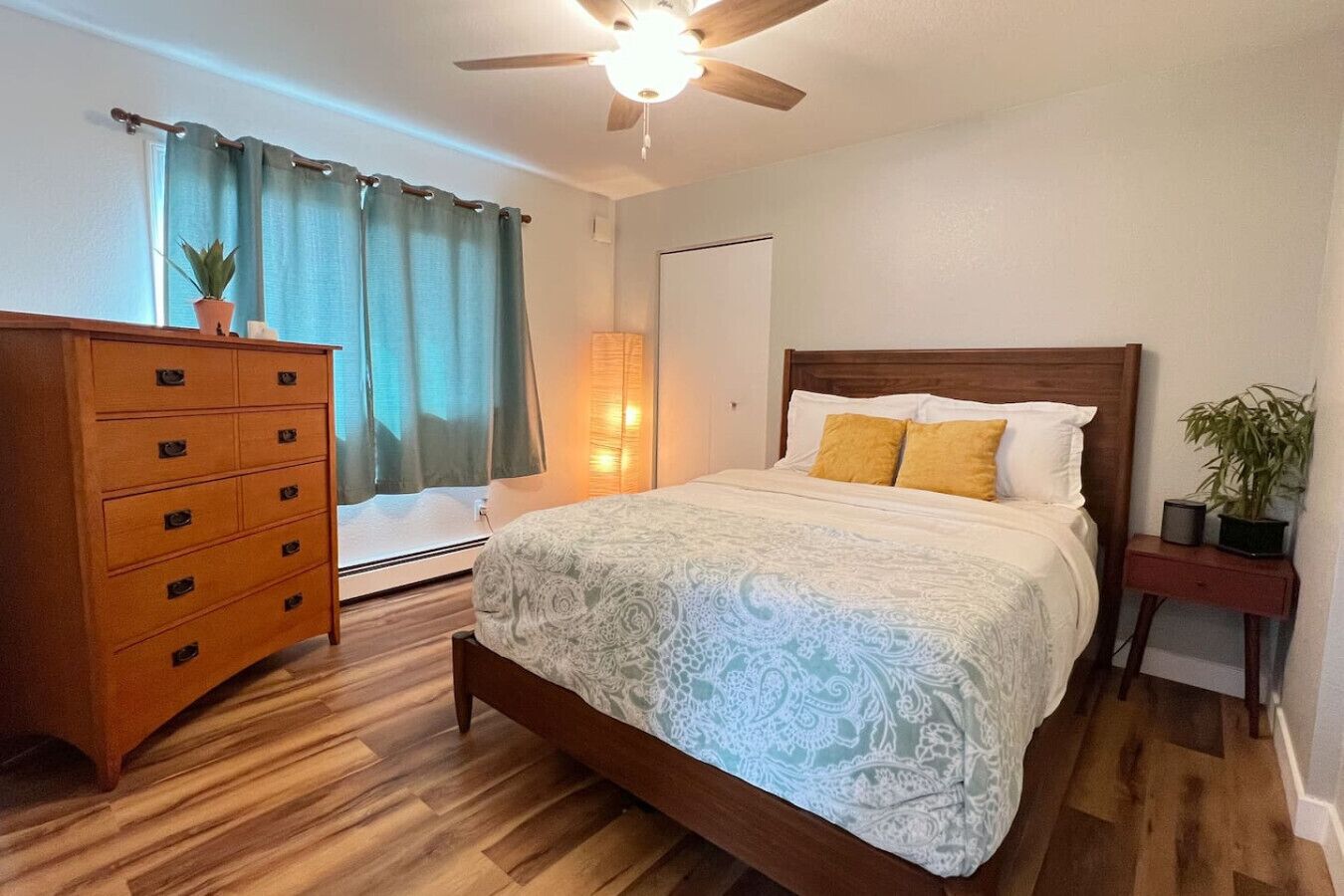 Un dormitorio con cama de madera, cómoda, ventilador de techo, ventana con cortinas y mesita de noche con una planta.