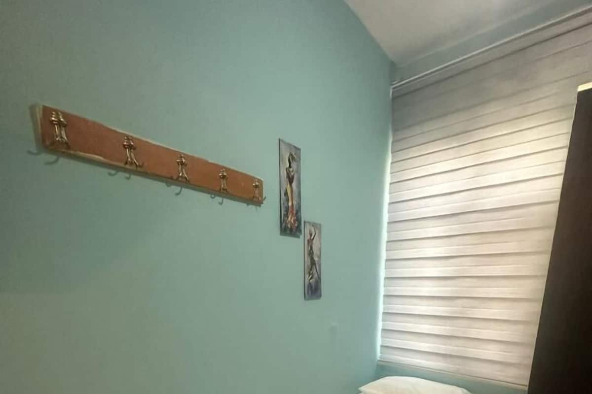 A small, single-bed room with a wooden headboard, a window with blinds, and a wall-mounted coat rack with a few items.