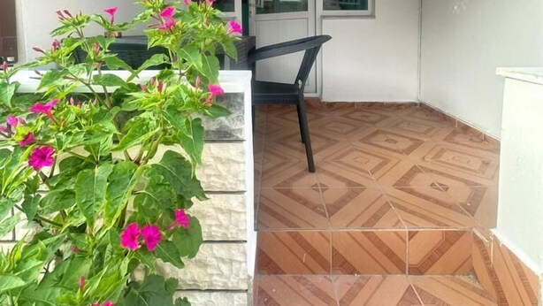 Een kamer met betegelde vloer, een raam en een deur. Er staat een stoel en een pot met roze bloemen.