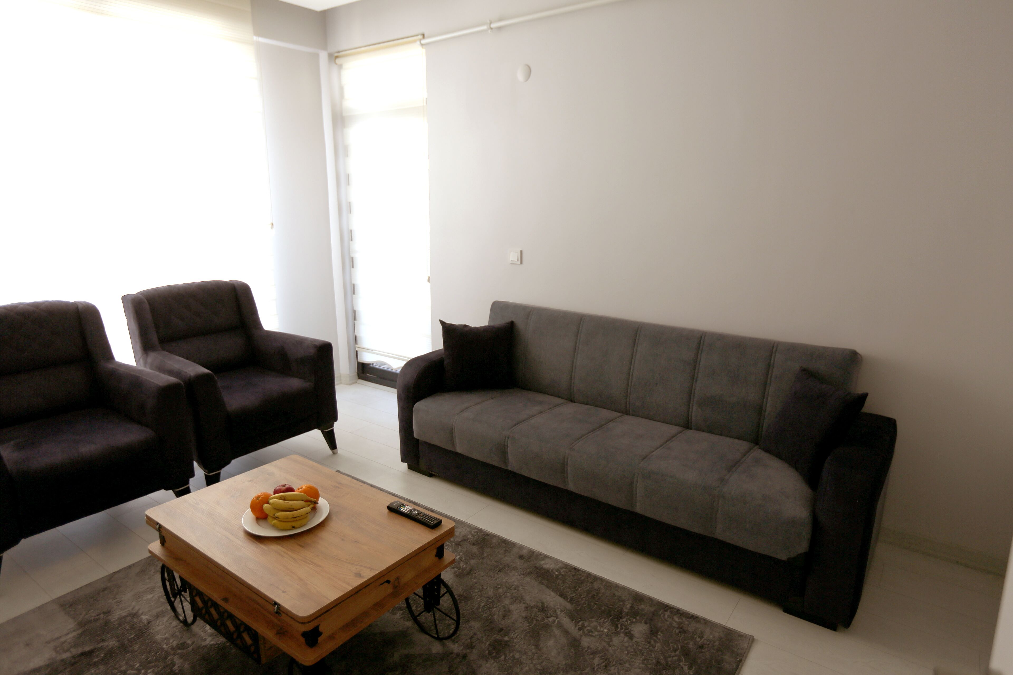 A living room with a grey sofa, two armchairs, a wooden coffee table with fruit and a remote control, and a grey carpet.