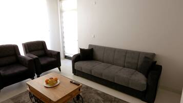 A living room with a grey sofa, two armchairs, a wooden coffee table with fruit and a remote control, and a grey carpet.