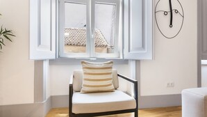 A room with a wooden floor, a white chair with striped cushions, a window with a view of buildings, and a wall-mounted abstract art piece. - Colosseum Building - Apartment Gamma (Lisbon)