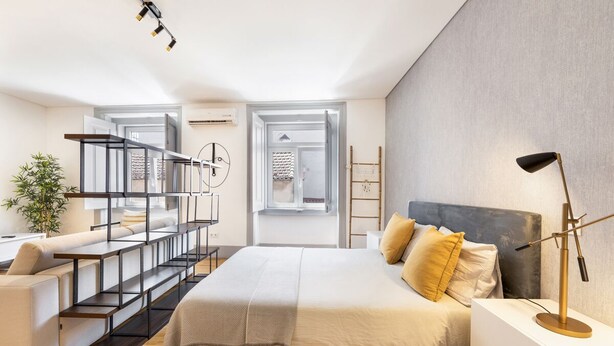 A modern bedroom with a large bed, a desk, and a chair. There is a plant and a ladder in the background. - Colosseum Building - Apartment Gamma (Lisbon)
