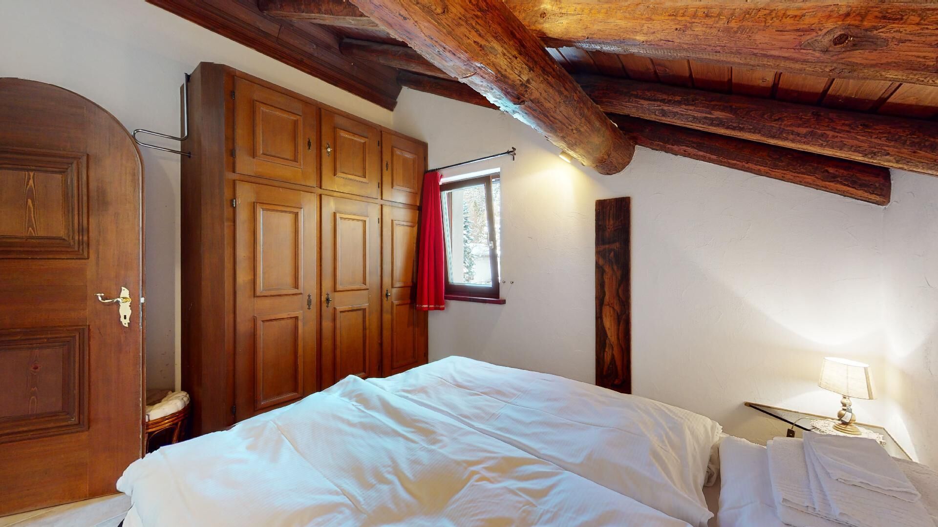 Une chambre avec une armoire en bois, un lit avec une literie blanche, une fenêtre avec un rideau rouge et un plafond avec une poutre en bois.