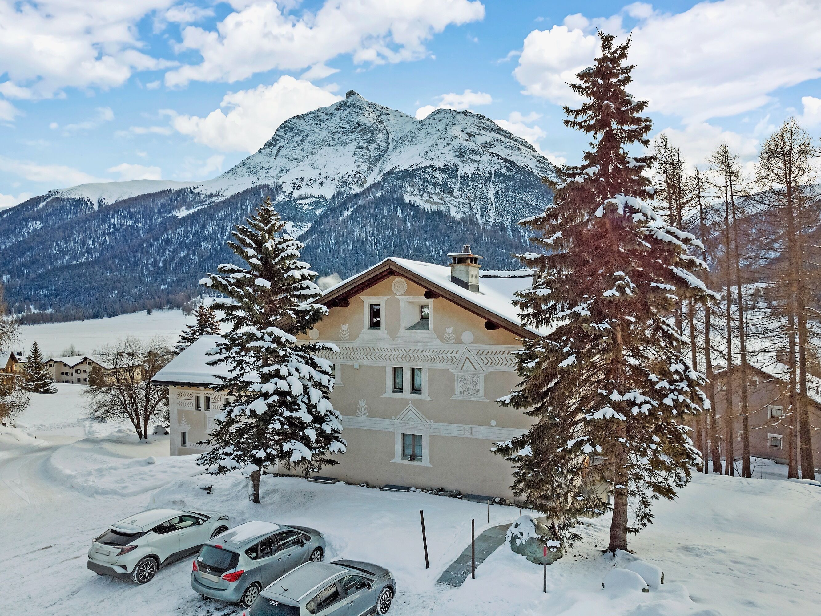 A snow-covered building with a mountain backdrop, parked cars, and snow-laden trees.