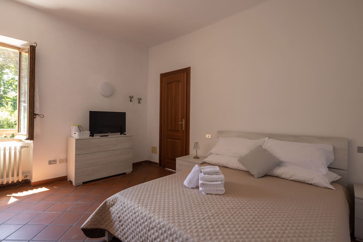 Um quarto com uma cama, uma televisão sobre um aparador, uma porta de madeira e uma janela.