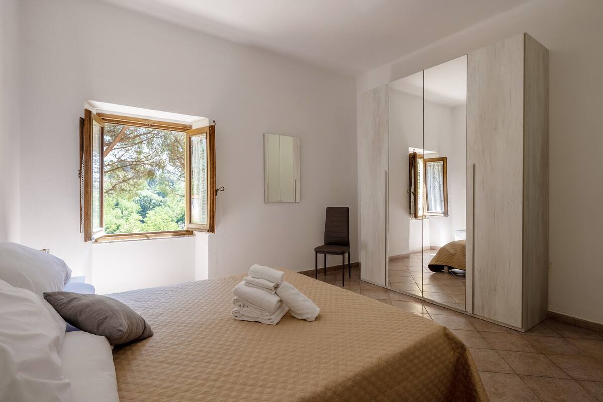 Quarto com cama, uma cadeira, guarda-roupa e janela com vista para árvores.