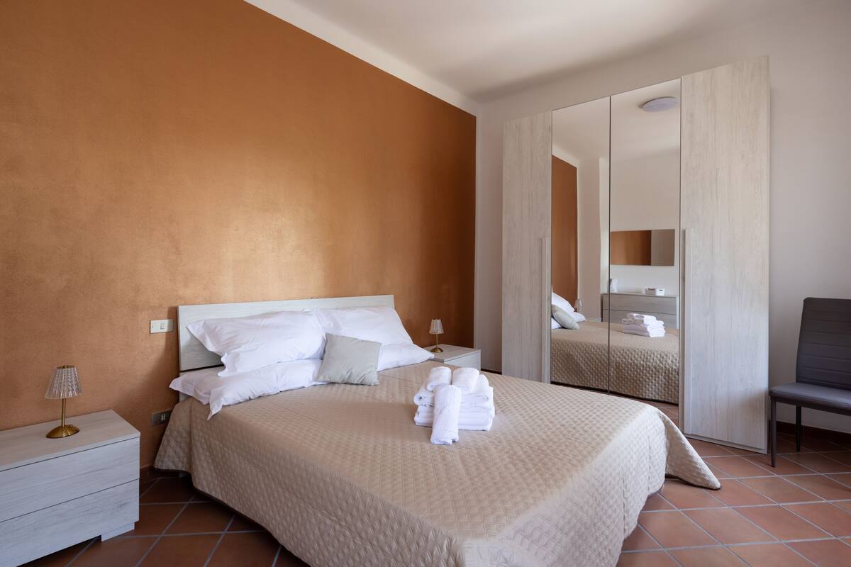 Cama bem arrumada com almofadas e toalha dobrada, mesa de cabeceira com abajur e uma cadeira em um quarto com espelho grande e guarda-roupa.