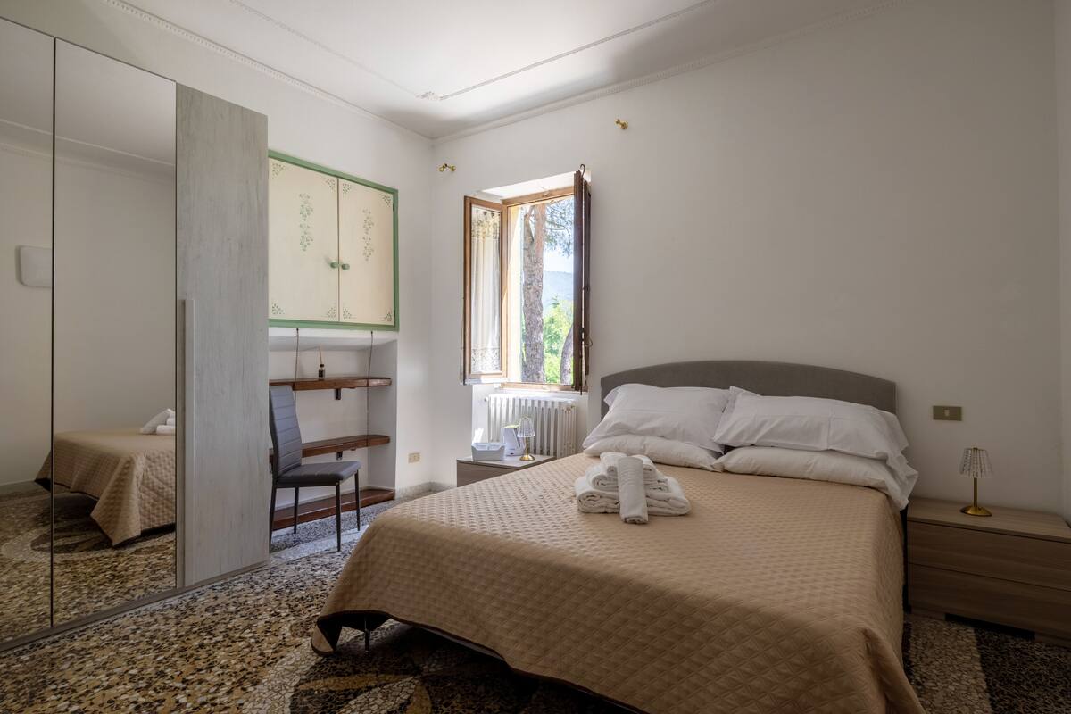Quarto com cama, cadeira, escrivaninha, espelho e janela.
