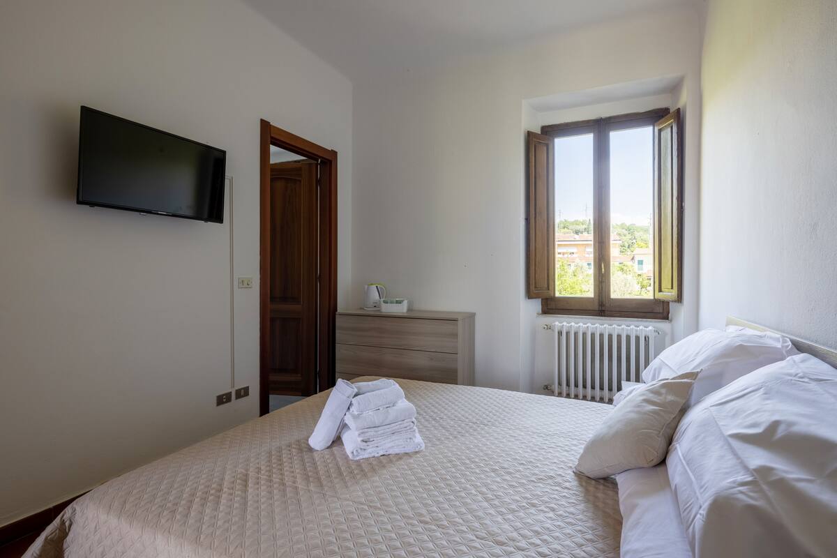 Quarto com cama, televisão na parede, cômoda e janela com vista para edifícios.