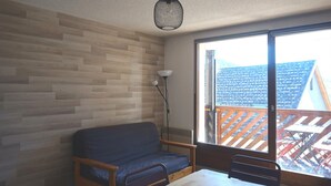 A room with a wooden wall, a blue sofa, a table, and a large window with a view of a roofed area. - Cozy apartment in Autrans center, 4 pers, south terrace, pets allowed (Autrans-Méaudre en Vercors-Autrans)