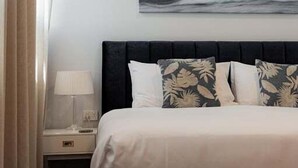 Ett hotellrum med en säng, ett nattduksbord, en lampa, en väggmonterad luftkonditioneringsanläggning och en tavla som föreställer en våg.