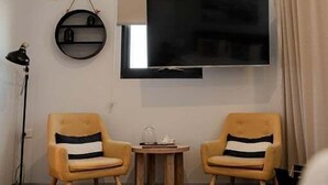 Ett hotellrum med en platt-TV som är monterad på väggen, två fåtöljer med randiga kuddar, ett litet träbord, en golvlampa och ett stort fönster med gardiner.