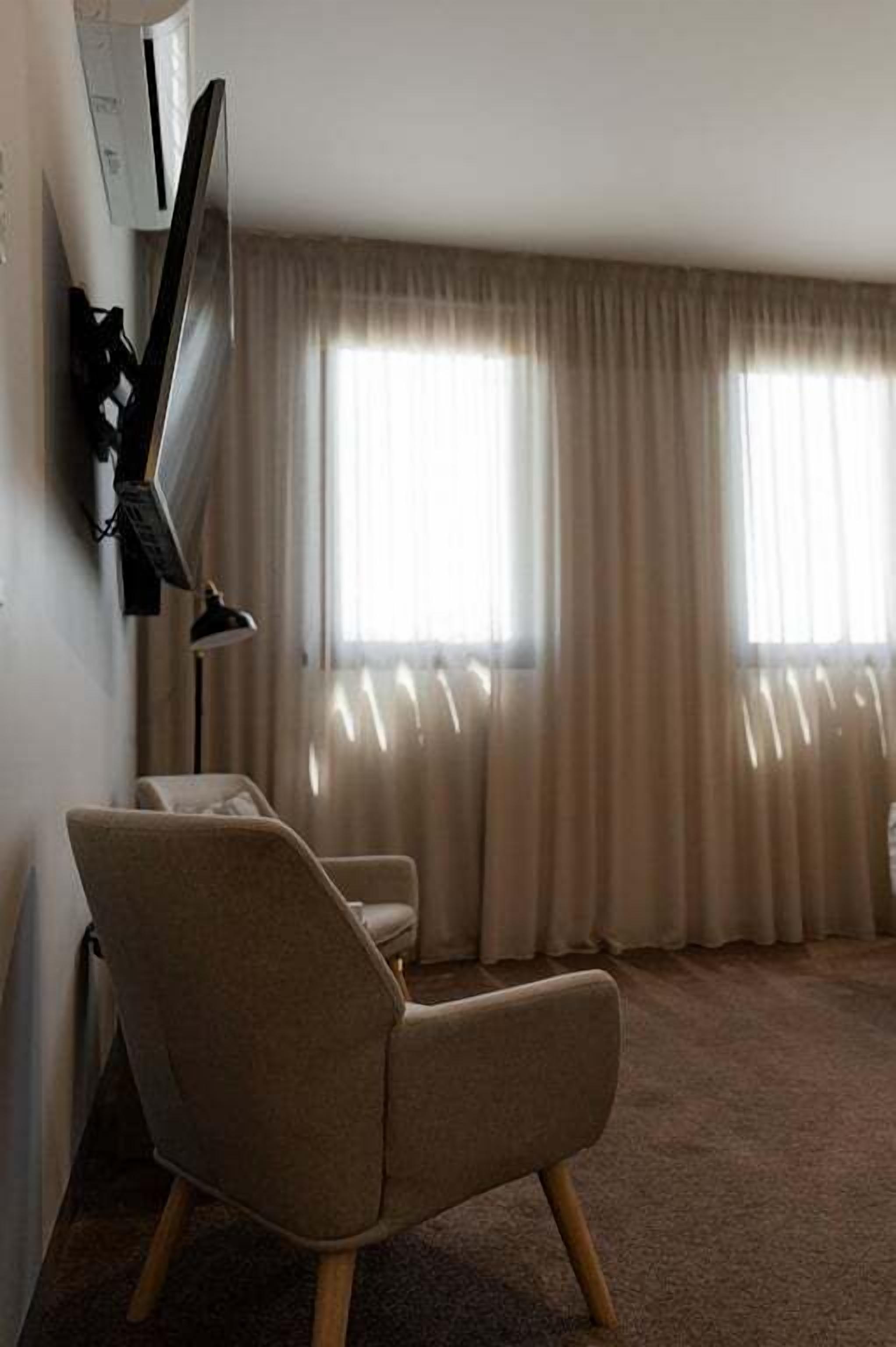Een kamer met een enkele beige fauteuil, een flatscreen televisie aan de muur en een groot raam met gordijnen.