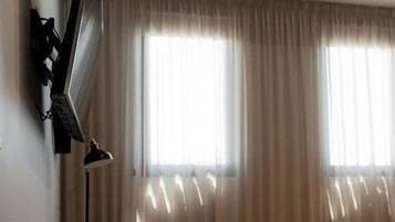 Een kamer met een enkele beige fauteuil, een flatscreen televisie aan de muur en een groot raam met gordijnen.