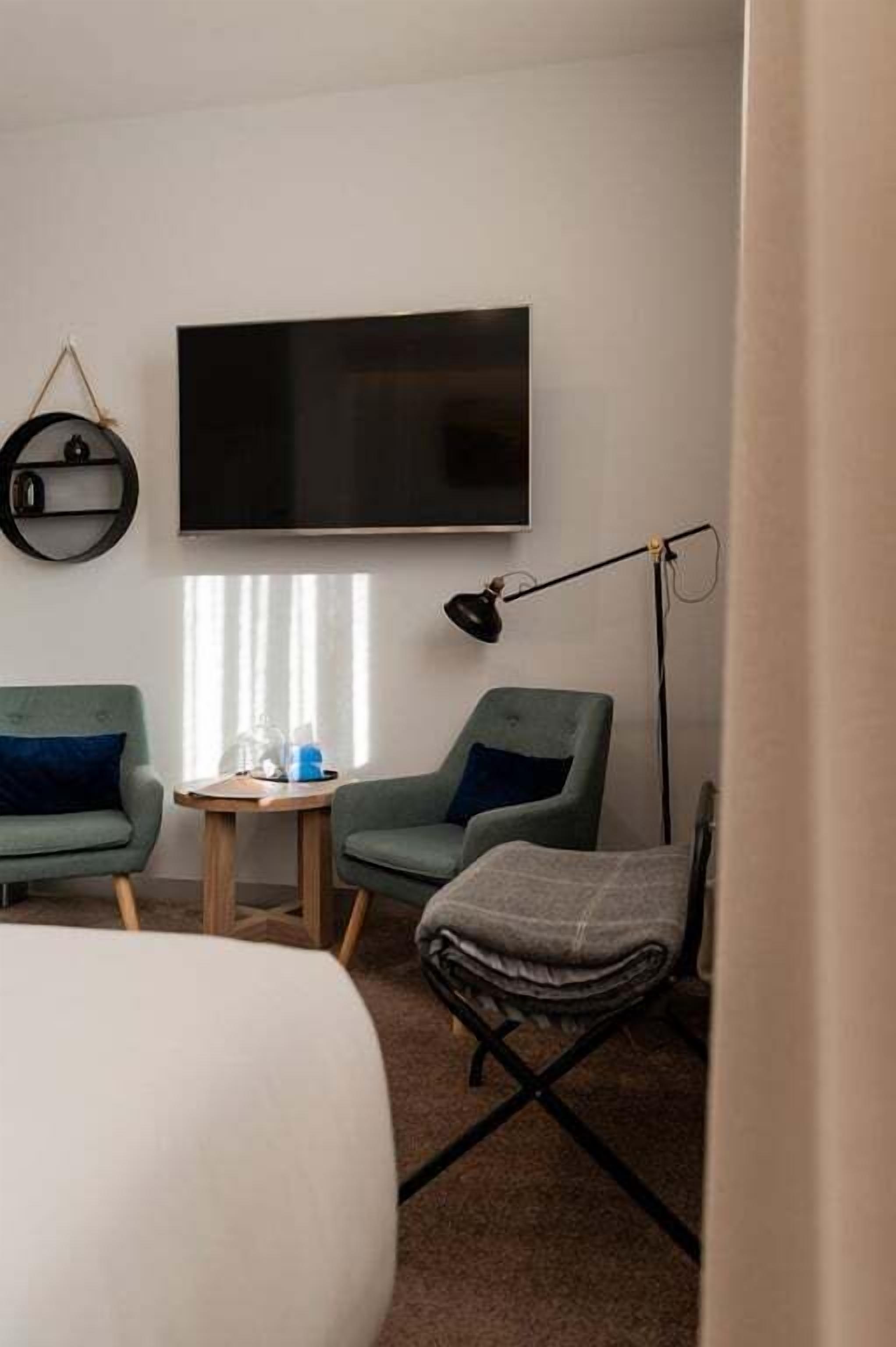 Una habitación de hotel con una cama, dos sillones verdes, una mesita de madera, una lámpara de pie y un televisor en la pared.
