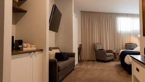 Habitación de hotel con sofá, sillón y cama. Tiene un televisor montado en la pared, una cafetera y una pequeña mesa con una silla.