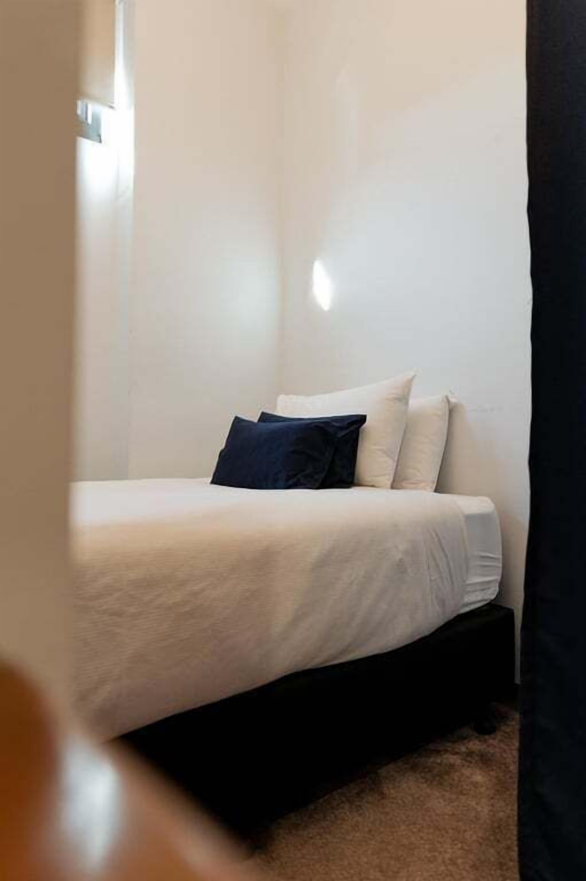A small, neatly made bed with a white mattress and two pillows, one dark and one light, in a corner of a room with white walls and a visible door frame.