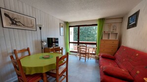A room with a dining table, chairs, a red sofa, a TV, and a window with curtains. - Cozy 3* apartment for 6, near slopes, with parking (Autrans-Méaudre en Vercors-Autrans)