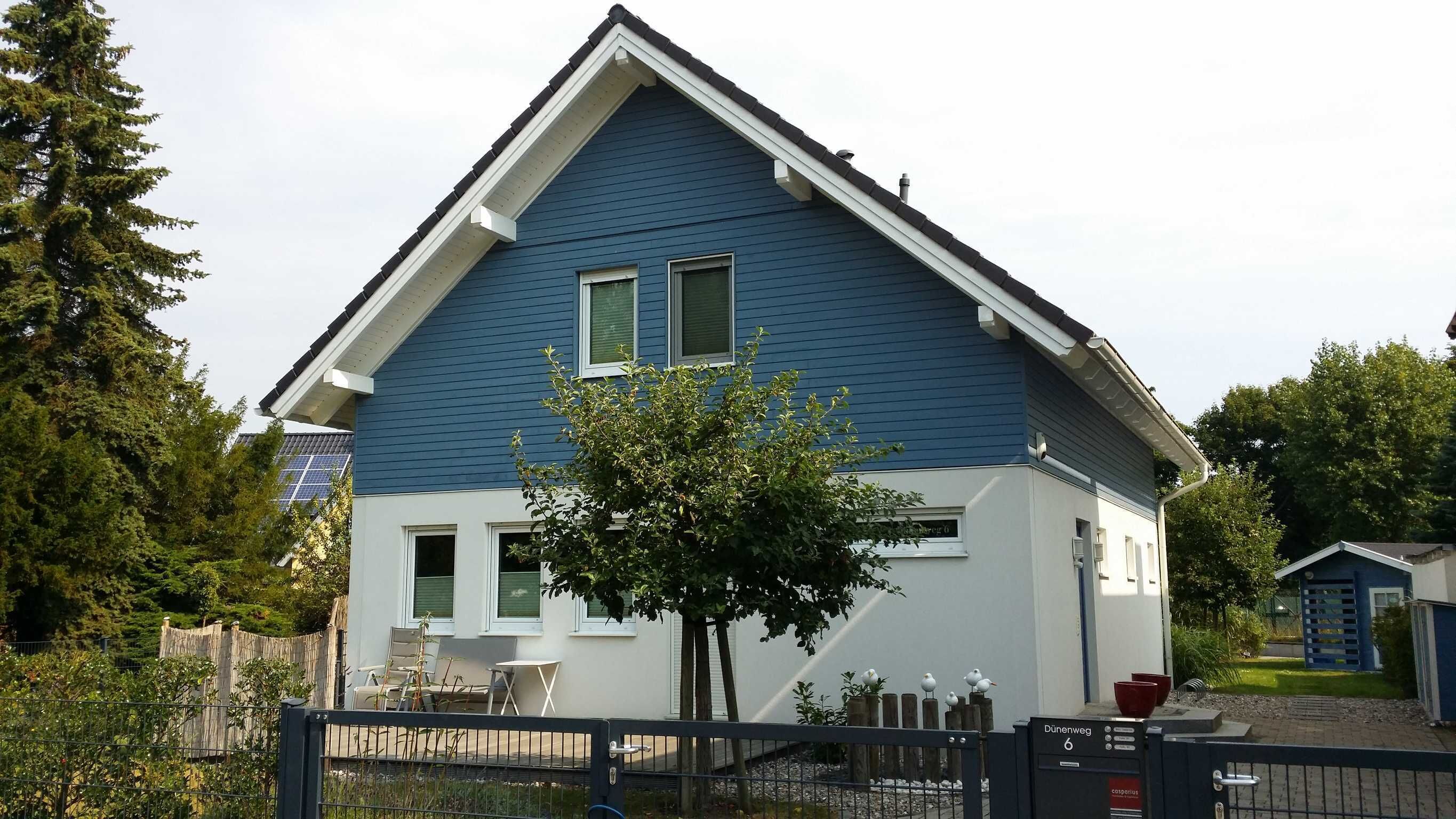 Ein zweistöckiges Haus mit blauer Fassade, einem kleinen Vorgarten mit Baum und einem eingezäunten Eingang.