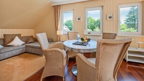 Un salon moderne avec un canapé d’angle, des fauteuils en osier, une table ronde et de grandes fenêtres donnant sur la verdure.