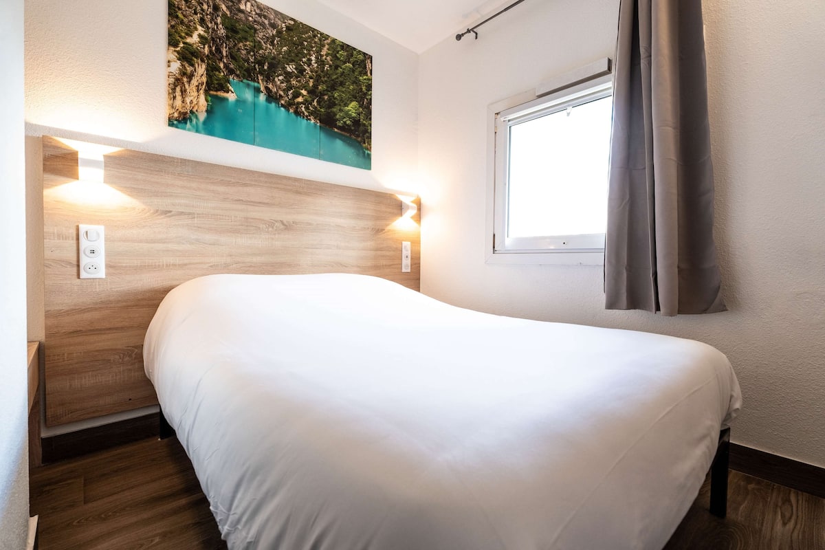 A neatly made bed in a room with a wooden headboard, a window with curtains, and a wall-mounted light fixture.