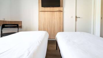A hotel room with two single beds, a wooden wardrobe, a mounted TV, and a chair.