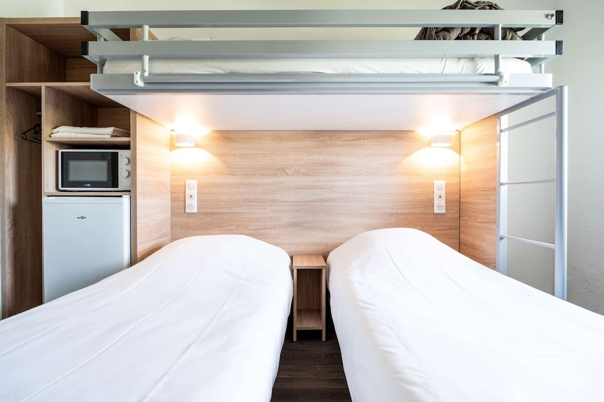 A compact room with two beds, a small kitchenette, and a wooden headboard.