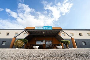 Entrada de un hotel con un cartel que dice "Kyriad DIRECT", asientos exteriores y cielo despejado.