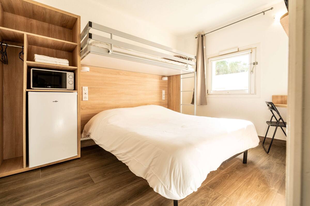 A compact room with a bunk bed, a small kitchenette with a microwave and refrigerator, and a window with curtains.