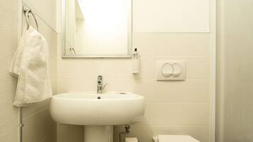Un baño con un lavabo blanco, una toalla colgada en un perchero, un espejo y un inodoro.