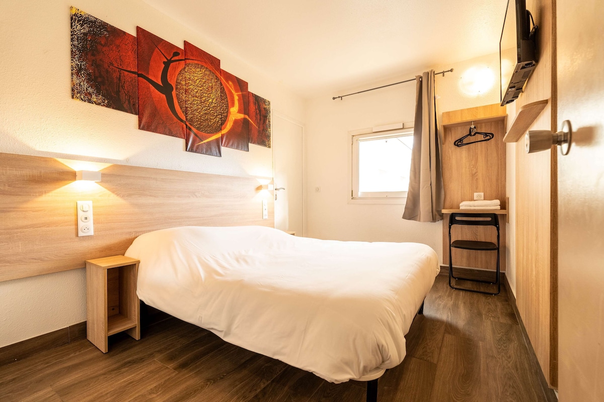 A modern hotel room with a wooden headboard, a bed with white bedding, a small bedside table, a wall-mounted TV, and a window with a curtain.