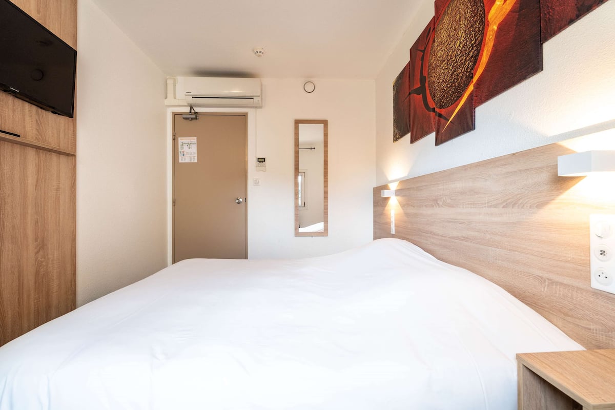 A hotel room with a bed, wooden headboard, wall-mounted artwork, a mirror, and an air conditioning unit.