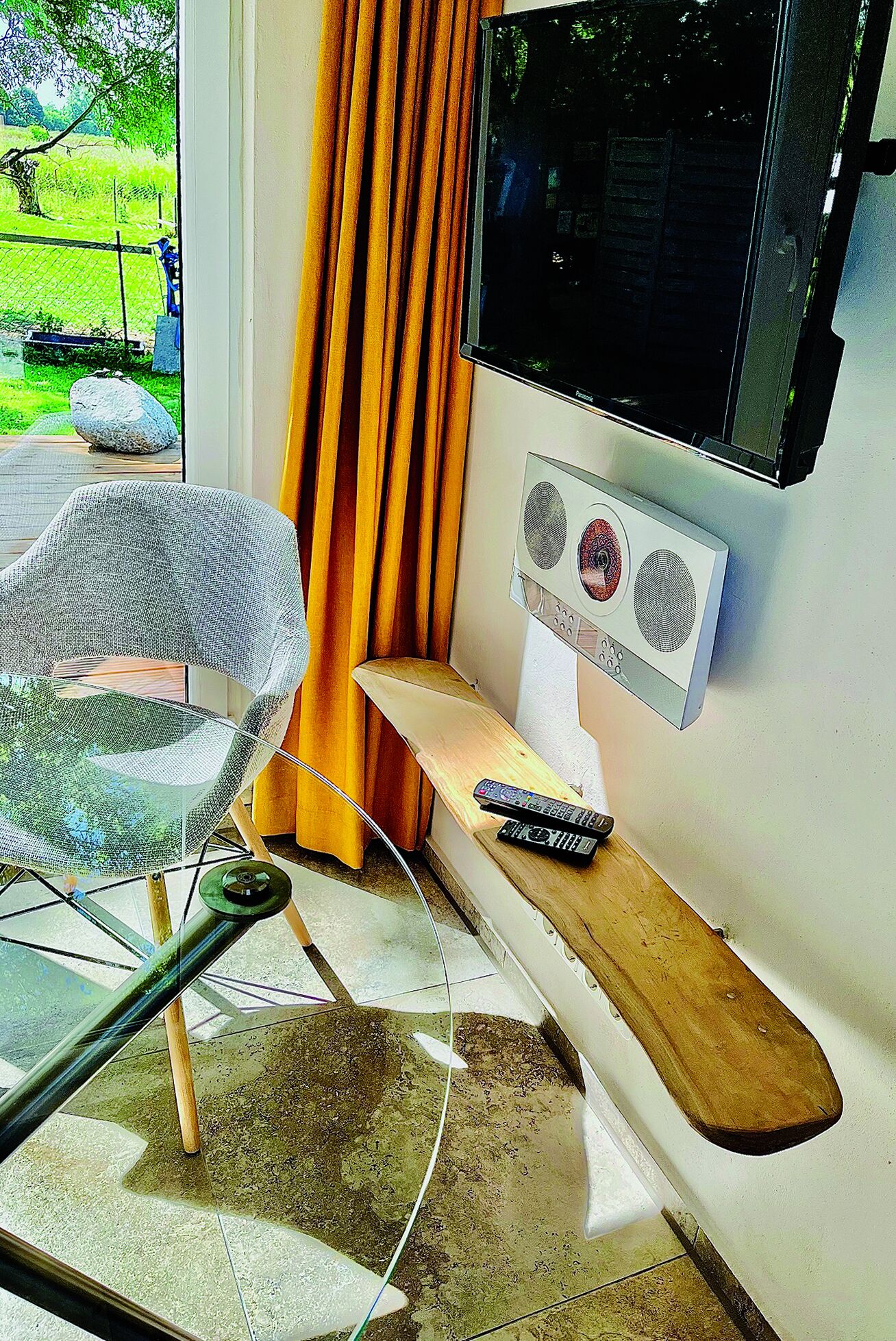 Een kamer met een glazen stoel, een houten plank met een afstandsbediening, een televisie en een raam met gordijnen.