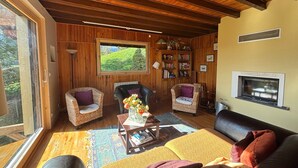 A cozy living room with wooden walls, a large window, a fireplace, and a bookshelf.
