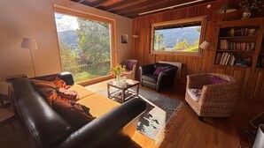 A cozy living room with a leather sofa, a wicker armchair, and a black leather chair. There are two large windows offering a view of the outdoors.